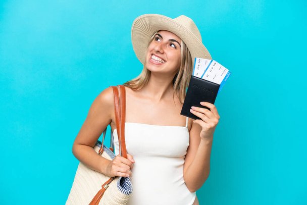 Giovane donna caucasica in costume da bagno in possesso di un passaporto isolato su sfondo blu guardando in alto mentre sorride - Foto, immagini
