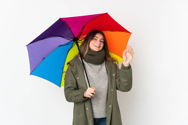 Joven mujer caucásica sosteniendo un paraguas aislado sobre fondo blanco saludando con la mano con expresión feliz - Foto, Imagen