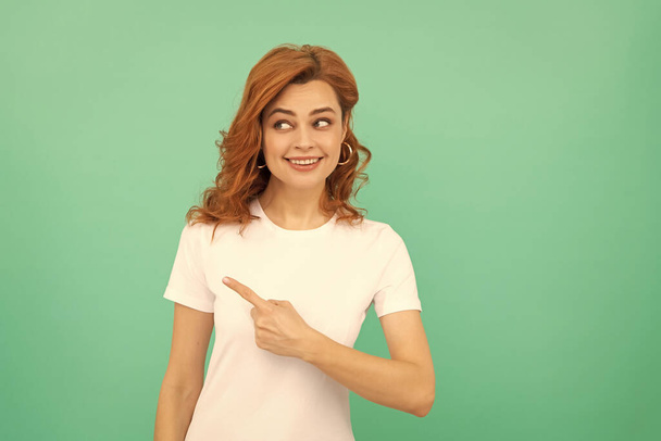 donna rossa sorridente con i capelli ricci che punta il dito su sfondo blu, spazio copia, pubblicità. - Foto, immagini