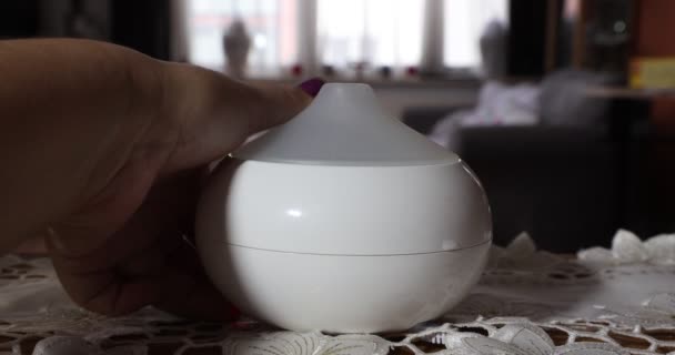 Female hands turning on a white scented aroma oil diffuser humidifier inside a home, against a blurred background, with a color changing lamp. Concept of ambient relaxation at home - Filmati, video