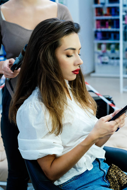 Güzel esmer bir kadın saçını güzellik salonunda şekillendirirken telefonu kullanır.. - Fotoğraf, Görsel