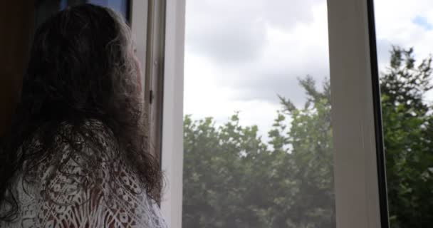 Mature woman looking out the window and getting fresh air on a cloudy morning, just showered, smiling happily and taking energy to start the day with enthusiasm, gray black loose long hair wet - Filmati, video