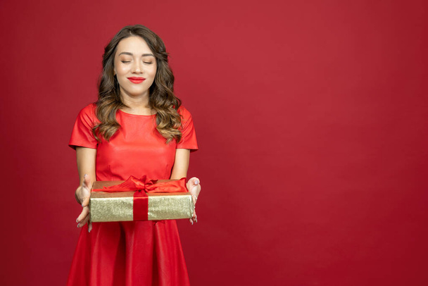 A beautiful brunette woman is looking at a gift and smiling pretty in a red dress on a red background with copy space. - Foto, imagen