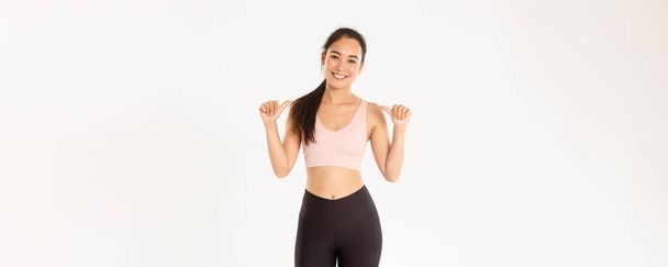 Sport, wellbeing and active lifestyle concept. Smiling slim and strong, attractive asian female fitness coach, personal instructor or trainer pointing at herself, your gym logo, white background. - Photo, Image