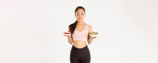 Active lifestyle, fitness and wellbeing concept. Portrait of tempting cute asian girl trying resist temptation as looking at delicious cake, being on diet, need eat healthy salad, licking lips. - Photo, Image