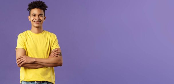 Close-up portrait of confident, smart and professional young male student with dreads, yellow t-shirt, cross arms over chest and smiling pleased, knows what he doing, purple background. - Photo, Image