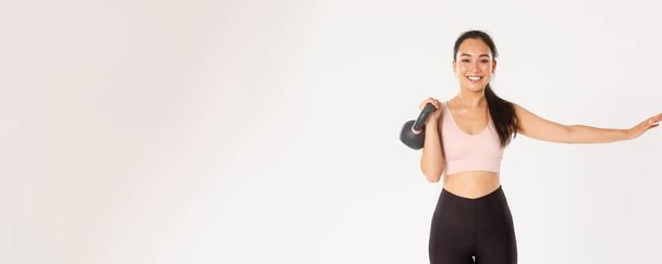 Sport, wellbeing and active lifestyle concept. Smiling beautiful asian fitness girl, gym coach extend one hand and lift kettlebell, bodybuilding, gaining muscle strength, standing white background. - 写真・画像