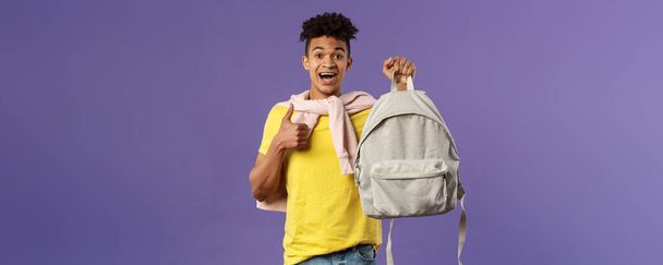 Portret pogodnego młodego przystojnego studenta, mężczyzna polecający plecak, trzymający plecak i kciuki do góry, kupił nowy sprzęt na semestr uniwersytecki, fioletowe tło. - Zdjęcie, obraz