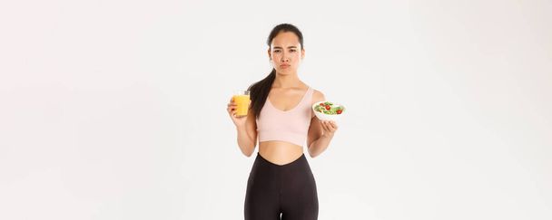 Sport, welzijn en actief lifestyle concept. mokken en terughoudend schattig aziatisch meisje haat eten gezond, met salade en sinaasappelsap met geïrriteerde pruilen gezicht, haat het zijn op dieet en workout. - Foto, afbeelding