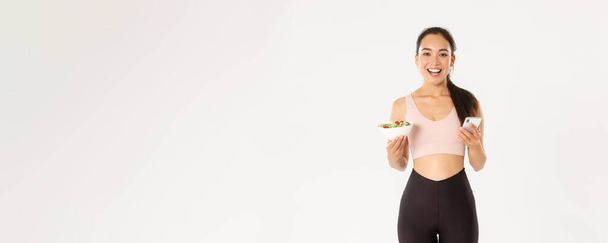 Sport, wellbeing and active lifestyle concept. Smiling slim asian fitness girl in sportswear, holding salad and mobile phone, using eating reminder app, diet control application, checking calories. - Фото, зображення