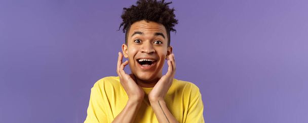 Close-up portrait of extremely happy, enthusiastic young man hear fantastic news, looking surprised and excited, touching face in joy, smiling upbeat look camera astonished, purple background. - Photo, image