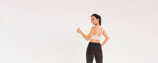Full length of motivated and satisfied smiling asian female coach, girl athelte in sportswear turn behind to show thumbs-up at camera, ready for productive fitness training, white background. - Valokuva, kuva
