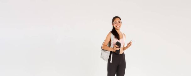 Full length of smiling healthy, slim asian girl going fitness training, female athelte carry backpack with workout equipment and water bottle, using mobile phone sports application, white background. - Photo, Image