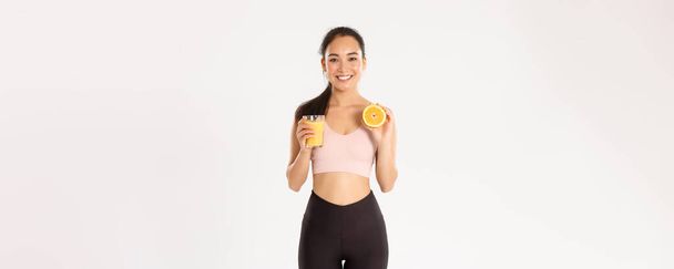 Sport, wellbeing and active lifestyle concept. Portrait of smiling healthy and slim asian girl advice eating healthy food for breakfast, gain energy for workout, hold fresh juice and orange. - Photo, Image