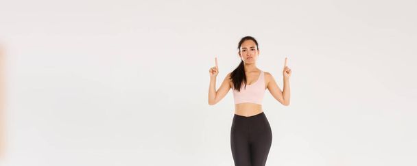 Sport, gym and healthy body concept. Full length of unamused and skeptical fitness girl, asian female athlete in sportswear complaining, sulking displeased and pointing fingers up, white background. - Photo, Image