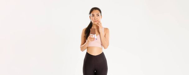 Sport, wellbeing and active lifestyle concept. Doubtful and indecisive asian fitness girl holding chocolate bar and thinking about eating it, looking away hesitant, worried about calories. - Zdjęcie, obraz