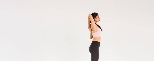 Full length profile of focused strong asian female coach, sportswoman doing fitness exercises, making hands stretching, lock arms on back, standing over white background. - Foto, Bild