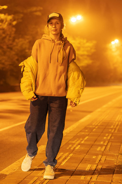 モダンな服装の女性は霧の道に沿って夜に荒れ果てた舗装に沿って歩きます. - 写真・画像