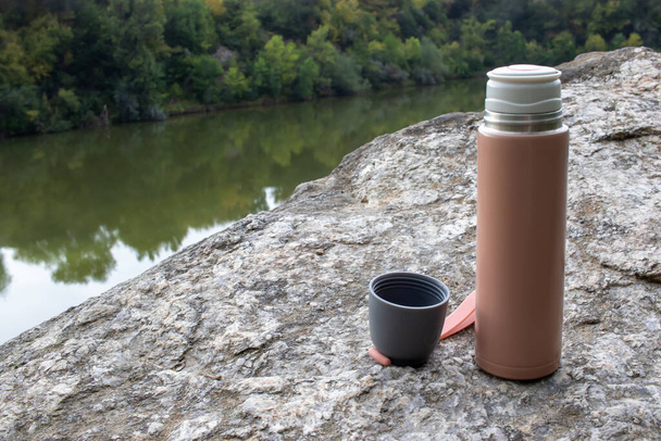 Beige thermos bottle and cup of tea or coffee on rock over the river. Beautiful summer, spring, autumn landscape. Picnic on the rock - Fotoğraf, Görsel