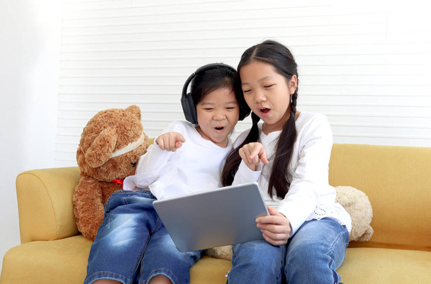 Two adorable sister girls hold digital tablet, put on headphones for online learning courses, pointing at screen, happy kids have fun study by using technology, children surfing internet. - Foto, Imagem