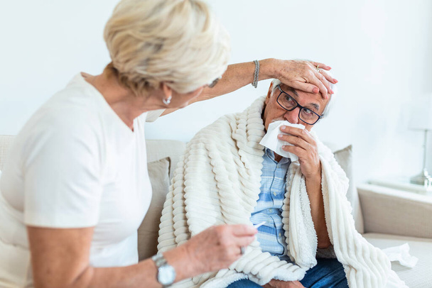 Пожилая женщина помогает больному мужу, когда они сидят на диване в гостиной. Старший с гриппом и бегающим носом. - Фото, изображение