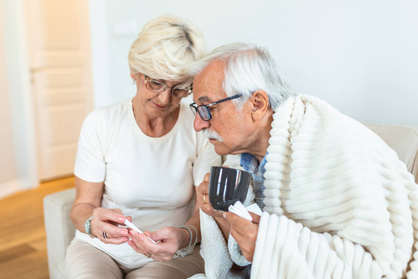 Sick senior man with cup of tea. His wife showing him a thermometer. Woman checking fever temperature of senior man sitting on bed. - Foto, Imagen