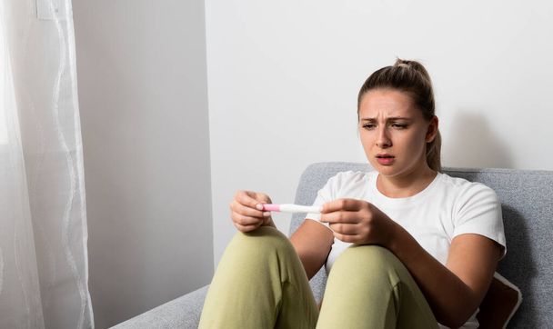 Young nervous woman is sitting at home holding a pregnancy test and is impatiently waiting for the test result. Maybe pregnant maybe not concept with anxious inpatient female on sofa in her apartment. - Photo, image