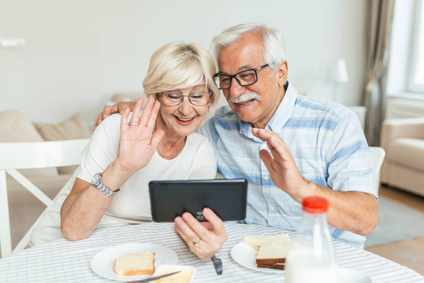 Happy older couple using computer tablet together at home, excited mature man and woman looking at mobile device screen, video call chatting online while having breakfast at home. - Fotografie, Obrázek
