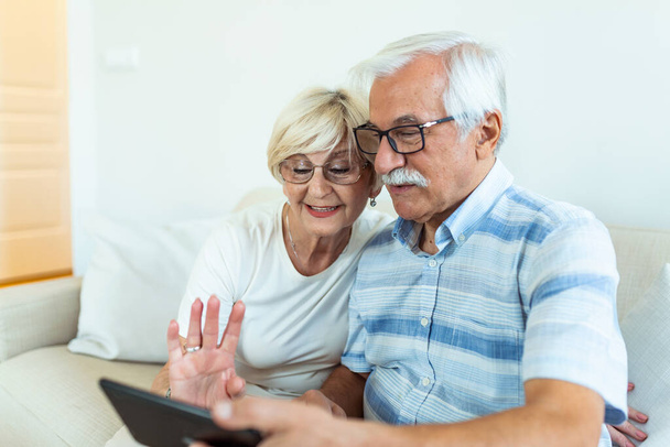 Couple of seniors smiling and looking at the same tablet hugged on the sofa - indoor, at home concept - caucasians mature and retired man and woman using technology - Foto, Bild