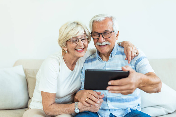 Couple of seniors smiling and looking at the same tablet hugged on the sofa - indoor, at home concept - caucasians mature and retired man and woman using technology - lockdown and quarantine lifestyle - Foto, imagen