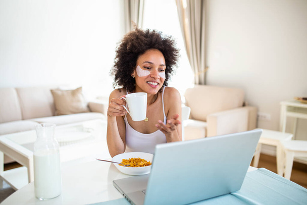 Гарна молода американка, яка друкує на ноутбуці вдома. Весела пані їсть кукурудзяні пластівці, п "є ранкову каву і працює на ноутбуку вдома. - Фото, зображення
