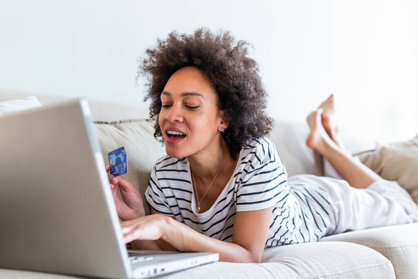 Интернет оплата. Счастливая жизнерадостная женщина смотрит на экран Интернета, совершая интернет-платеж. Счастливая молодая женщина с кредитной картой и ноутбуком сидит дома на диване. Онлайн-оплата - Фото, изображение
