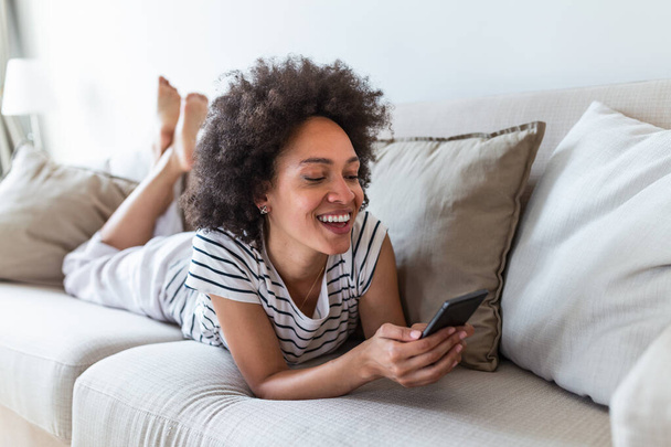 Pretty girl using her smartphone on couch at home in the living room. Young beautiful woman texting on her mobile phone while sitting at home in comfortable sofa - Zdjęcie, obraz