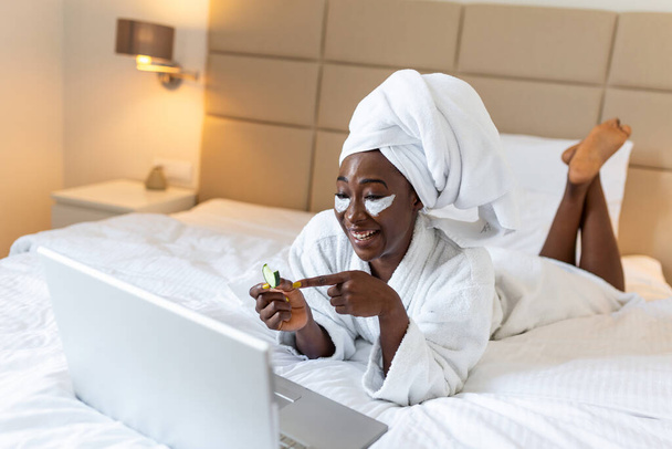 Wellness from home while working on laptop. Woman with cucumber for beauty treatment talking to her friends via video call on her laptop, laying ih bed in bathrobe and towel - 写真・画像