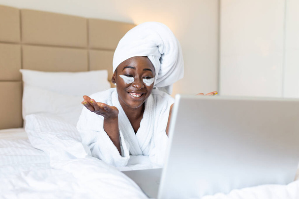 Beautiful young African woman in bathrobe resting on bed after shower video calling her friends on her laptop in the morning.Looking at laptop computer and talking to her friends via video call. - Foto, imagen