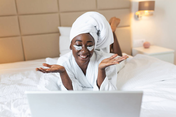 Young woman sitting at home with face beauty mask in front of her laptop. Cozy time at home. Self time and home spa concept. Beautiful African woman in bathrobe video calling her friends on her laptop - Foto, imagen