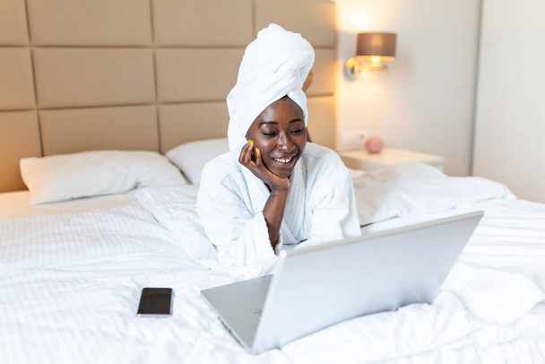 Улыбающаяся африканская молодая женщина лежит на кровати в халате и полотенце на голове с помощью ноутбука на кровати дома - Фото, изображение