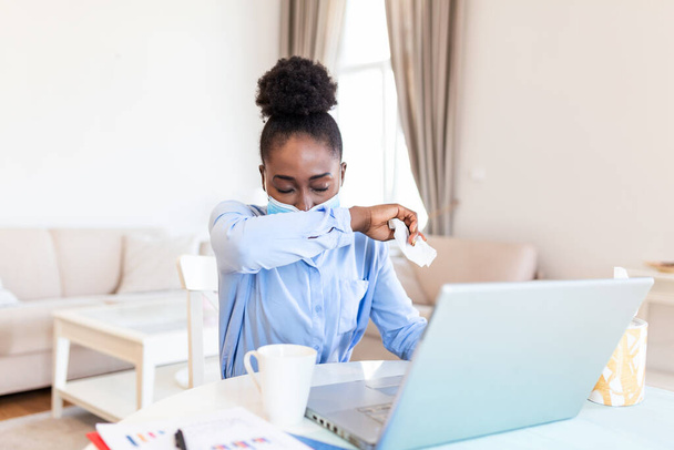 Young African businesswoman coughing into elbow in the home office. Businesswoman working in her home office while suffering from allergies. Women are sneezing and are cold during COVID-19 pandemic. - Zdjęcie, obraz
