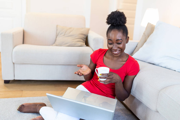 Attractive happy young african american woman sitting at the floor, using laptop computer, having video chat and drinking coffee - Fotografie, Obrázek