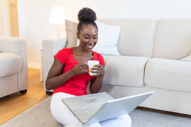 Αφροαμερικανή νεαρή γυναίκα που κάνει βιντεοκλήση με φίλους ή οικογένεια. Κρατώντας φλιτζάνι καφέ ή τσάι. Καθισμένος στο πάτωμα του σαλονιού. Όμορφη γυναίκα που νιώθει ευτυχισμένη - Φωτογραφία, εικόνα