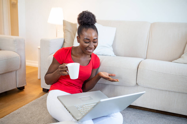 African american young woman having video call with friends or family. Holding cup of coffee or tea. sitting on the floor of living room. Beautiful woman feeling happy - Photo, image