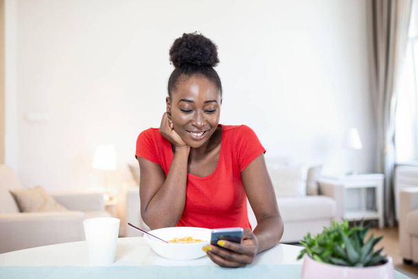 Young smilling woman sitting on table and enjoying first morning coffee or tea. Young amazing woman sitting indoors at the table with mobile phone. Looking at smart phone and texting her friends - Photo, Image