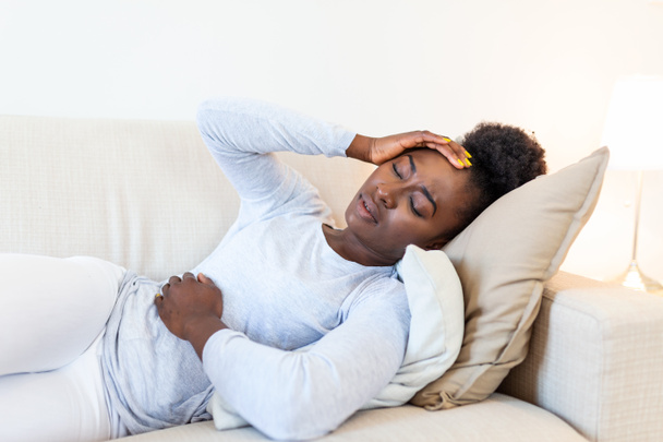 Portrait of a young black girl sitting on the couch at home with a headache and stomach pain. Beautiful woman suffering from chronic daily headaches. Sad woman holding her head because sinus pain - Valokuva, kuva