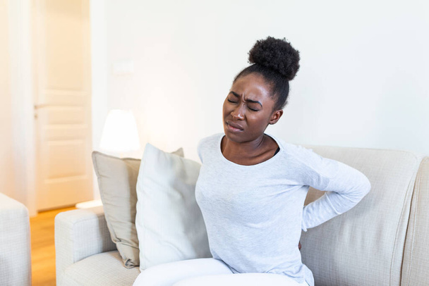 Young Black woman suffering from backache at home. Portrait of a young brunette girl sitting on the couch at home with a headache and back pain. Beautiful woman Having Spinal Or Kidney Pain - Photo, Image
