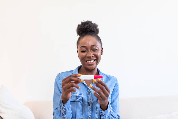 Young woman looking at pregnance test in happiness. Finally pregnant. Attractive black women looking at pregnancy test and smiling while sitting on the sofa at home - Fotografie, Obrázek