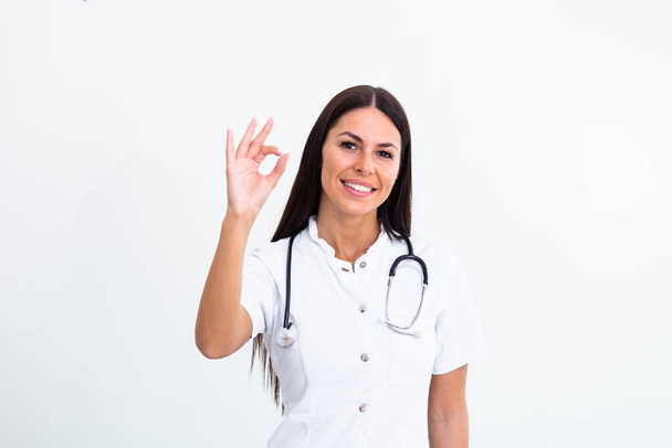 Ιατρική έννοια του νεαρού όμορφη γυναίκα γιατρός σε λευκό παλτό με φωνοενδοσκόπιο δείχνει ok σημάδι. Γυναίκα εργαζόμενος νοσοκομείο κοιτάζοντας κάμερα και χαμογελά, στούντιο, λευκό φόντο - Φωτογραφία, εικόνα