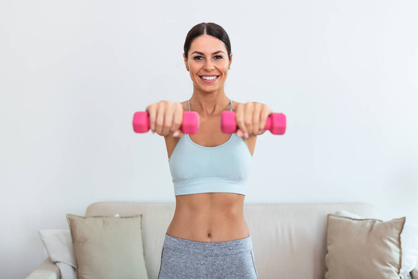 Woman exercising with dumbbells at home. Sporty beautiful woman exercising at home to stay fit. Young woman exercising at home in a living room. Fitness, workout, healthy living and diet concept. - Foto, imagen