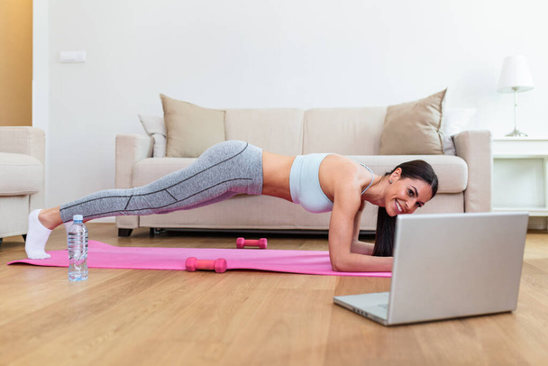 Young woman exercising at home in a living room. Video lesson. Young woman repeating exercises while watching online workout session. Beautiful young woman doing fitness exercise at home - Photo, Image