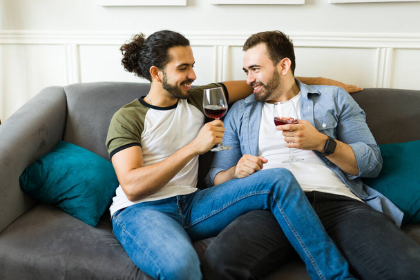 Beautiful gay couple smiling and looking happy during a romantic anniversary date at home drinking wine together on the sofa - Photo, image