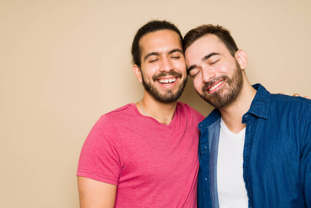 Χαλαρός καυκάσιος άντρας και ο χαρούμενος γκέι σύντροφός του αγκαλιάζουν και κλείνουν τα μάτια τους ενώ γελούν σε φόντο στούντιο. - Φωτογραφία, εικόνα